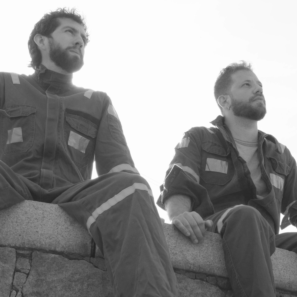 Two men in worn work overalls sitting on a stone wall against a bright sky, in black and white